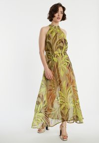 Robe longue en tissu transparent avec un motif de feuilles vertes et brunes. Encolure halter, taille cintrée et ourlet fluide. Associée à des talons dorés.