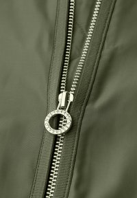 Primo piano di una giacca verde scuro parzialmente sbottonata con una cerniera rotonda in argento inciso con del testo.