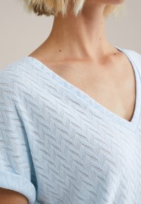 Gaiši zils adīts tops ar zīda rakstu, ar V formas izgriezumu un īsām piedurknēm. Audums ir mīksts uz tausti.