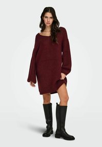 Bordowa dzianinowa sukienka swetrowa z szerokim dekoltem i długimi rękawami; posiada boczne rozcięcie, uzupełniona czarnymi kozakami za kolano.