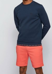 Uomo che indossa una camicia a maniche lunghe blu navy e pantaloni corti corallo, entrambi con dettagli ripetuti del logo "BOSS" sui bordi e sul petto.