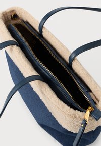 Tote din pânză bleumarin cu margini din faux shearling în nuanță de bej deschis. Prezintă două curele din piele și un fermoar aurit pentru compartimentul principal.