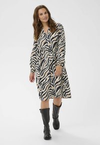 Šaty košilového typu v krémové, černé a béžové zebrované vzoru; dlouhé rukávy, výstřih do V, stažený pas, doplněné černými kotníkovými botami.