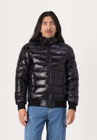 Zwarte pufferjas met glanzende afwerking, featuring horizontaal gestikte segments, een hoge kraag en elastische manchetten. Een klein logo is zichtbaar op de borst.