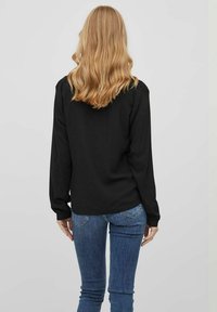 Camicetta nera a maniche lunghe con una texture liscia, caratterizzata da un taglio dritto e un dettaglio sulla spalla posteriore, abbinata a jeans blu aderenti.