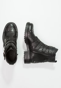 Pár černých kožených kotníkových bot s kovovými hroty, přezkami, postranním zipem a texturovanou podešví na bílém pozadí.