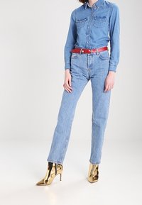 Jeansskjorta, blå med två bröstfickor; blåa jeans med hög midja; rött bälte; gyllene höga klackar med spetsiga tår och glansig yta.