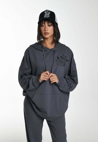 Sudadera gris de gran tamaño con un bolsillo frontal y detalles gráficos de flores en las mangas, combinada con joggers gris a juego. Gorra negra con diseño.