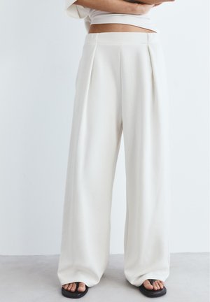 Žena má na sebe biele široké nohavice s vysokým pásom a záhybmi, v kombinácii s čiernymi remienkovými sandálmi, stojace pred jednoliatym bielym pozadím.