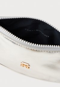 Srebrna metaliczna saszetka o gładkiej fakturze, z zamknięciem na zamek błyskawiczny i złotym akcentem w postaci logo. Wnętrze jest czarne z widocznym logo.