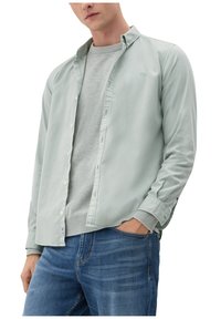Homme portant une chemise vert clair ouverte sur un pull gris avec un jean bleu, main dans la poche, sur fond blanc.