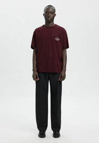 Burgundska majica iz mehke bombažne tkanine s kratkimi rokavi in potiskom na prsih z napisom "LA FLAMME ROUGE", v kombinaciji z ohlapnimi črnimi hlačami in črnimi čevlji.