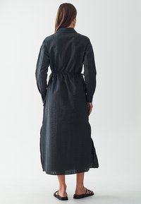 Abito lungo nero con superficie testurizzata, dotato di maniche lunghe, vita sfrangiata e spacchi laterali, realizzato in un tessuto leggero.