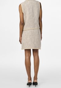 Dzianinowa sukienka bez rękawów w odcieniu beżowym z teksturowaną powierzchnią i kontrastowym wykończeniem. Długość do połowy uda, zestawiona z czarnymi szpilkami.