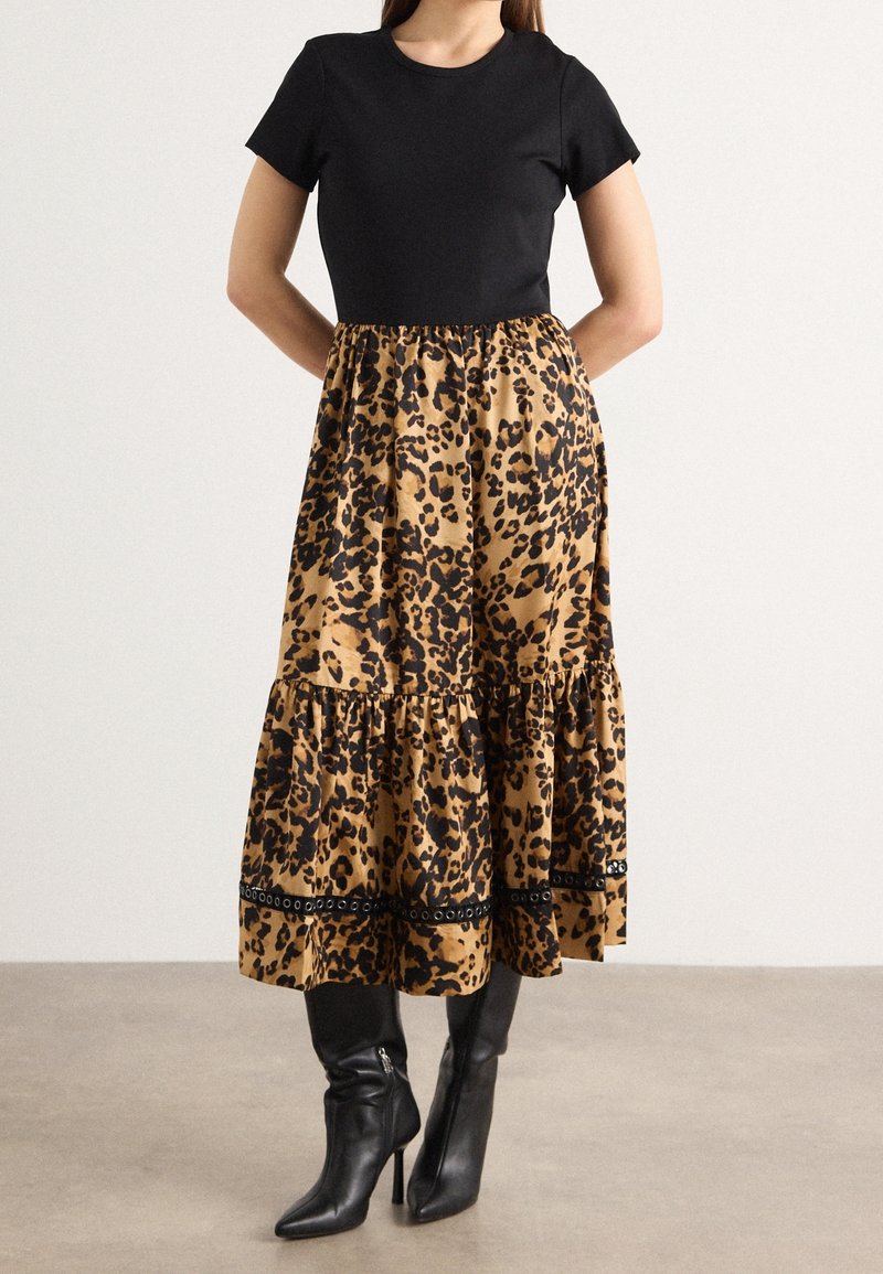 Femme portant un haut noir à manches courtes et une jupe midi imprimé léopard avec des bottes noires à talons hauts, debout à l'intérieur.