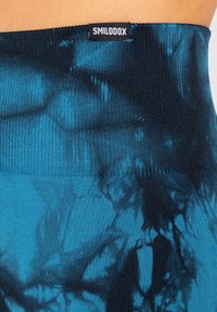 Zbliżenie na niebiesko-czarny pas materiału w technice tie-dye z prążkowaną fakturą i naszywką "SMILODOX" wszytą na środku.