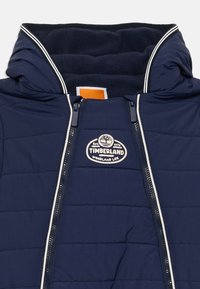 Marinblå Timberland vadderad jacka med vit kant, dubbla dragkedjor och krage fodrad med fleece, med Timberland-logotyp på bröstet.