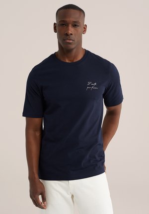 Muž oblečený v námornícky modrom tričku s bielym nápisom na hrudi a bielych nohaviciach, stojaci pred jednoduchým béžovým pozadím.