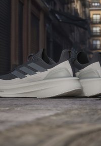 Ζευγάρι γκρι και λευκά αθλητικά παπούτσια Adidas Ultraboost τοποθετημένα σε λιθόστρωτο με αστικά κτίρια στο παρασκήνιο.