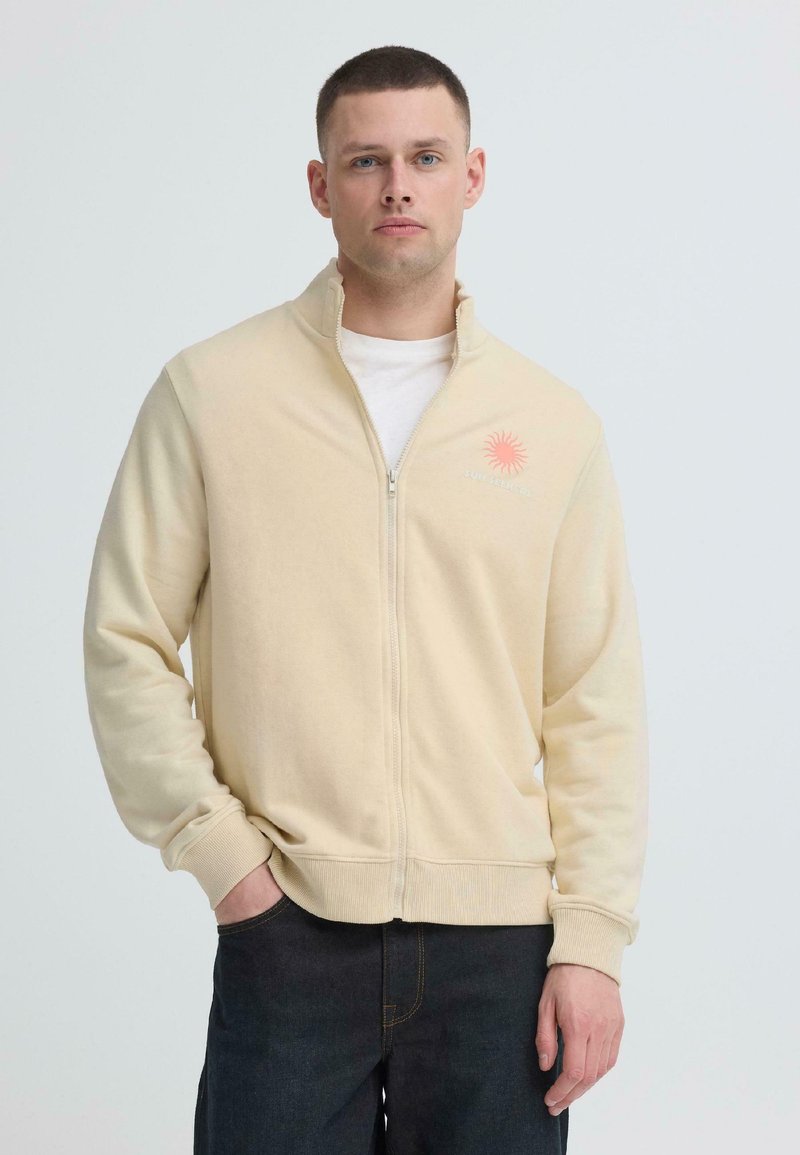 Homme portant une veste beige zippée sur une chemise blanche et un jean foncé, debout avec une main dans la poche devant un fond uni.