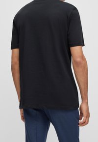 Man in een eenvoudig zwart T-shirt met korte mouwen en blauwe broek, van achteren gezien, tegen een effen lichtgrijze achtergrond.