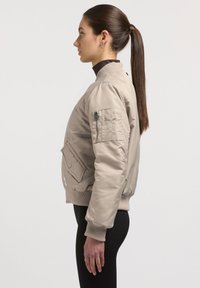 Beige Bomberjacke mit gerippten Bündchen, einer Vordertasche und einer Reißverschlusstasche am Arm. Glatte Textur, taillierte Passform und Stehkragen.