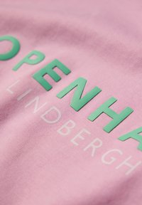 Vaaleanpunainen puuvillakangas, jossa on kohotetut vihreät kirjaimet, jotka kirjoittavat "COPENHAGEN" ja vaaleanharmaat "LINDBERGH." Sujuva pinta, jossa on hienoinen kiilto.