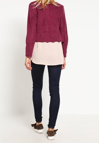 Burgundy mockaskinnjacka med en kort design lager ovanpå en ljusrosa topp, matchad med mörka skinny jeans och bruna slip-on skor.