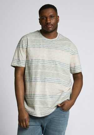 Man staat met één hand in zijn zak, draagt een lichtgekleurde gestreepte T-shirt en blauwe spijkerbroek, effen grijze achtergrond.