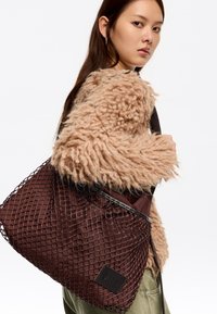 Brązowa siatkowa torba na ramię o teksturowanej powierzchni, z solidnym spodem i czarną naszywką z logo. Torba posiada wytrzymałe czarne uchwyty do noszenia.