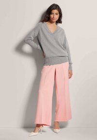 Lichtgrijze V-halstrui, relaxte pasvorm, gecombineerd met wijde, lichtroze broek. Model draagt witte puntige hakken. Glad stoffen texturen.