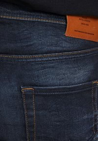 Jeans en denim bleu foncé avec une surface texturée, dotés de coutures orange, d'une étiquette en cuir au niveau de la taille et de poches arrière avec des ouvertures standard.