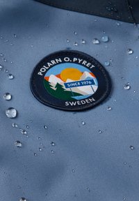 Sininen kangas, jossa on vesipisaroita, sisältäen pyöreän kirjotun tarran, jossa on vuoria, aurinko ja teksti "POLARN O. PYRET SWEDEN."