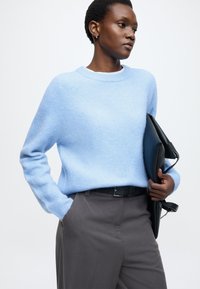 Světle modrý svetr z měkkého materiálu s žebrovanými manžetami, kombinovaný s šedými kalhotami a černou kabelkou drženou v levé ruce.