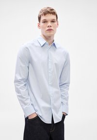 Camisa azul claro con botones, con sutiles rayas blancas verticales, mangas largas y cuello clásico. Usada de manera informal con jeans oscuros.