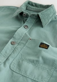 Hellgrüne Hemdjacke mit einem Cordkragen, ausgestattet mit metallischen Knöpfen und einer Brusttasche mit einem schwarzen Etikett. Weicher Stoff.