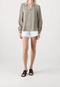 Kvinna som bär en långärmad olivgrön skjorta med krage, vita slitna denimshorts och svarta öppna klackskor står framför en enfärgad bakgrund.