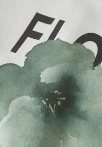 Baumwollstoff mit einem grünen, aquarellierten Blumenmuster, das weiche, sanfte Texturen und teilweise sichtbare, kräftige schwarze Schriftzüge zeigt.