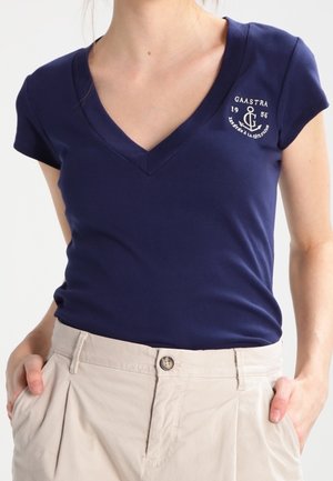 Femme portant un t-shirt à col en V bleu marine avec un logo d'ancre blanc et du texte sur la poitrine, assorti à un pantalon beige taille haute.