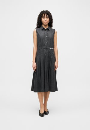 Αμάνικο τζιν φόρεμα σε σκούρο γκρι με γιακά, κουμπιά μπροστά, εφαρμοστή μέση και πλισέ φούστα. Μαύρα φλατ παπούτσια. Απλός σχεδιασμός.