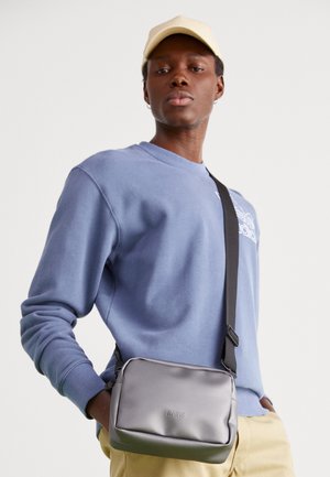 Persona joven con gorra beige, sudadera azul, pantalones amarillos y bolso cruzado gris de RAINS con correa negra.