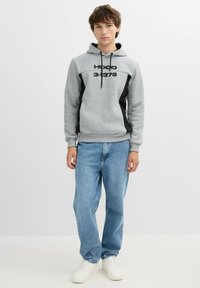 Sudadera gris con detalles negros y texto impreso. Combinada con jeans de color azul claro y zapatillas blancas. Diseño sencillo con capucha ajustable.