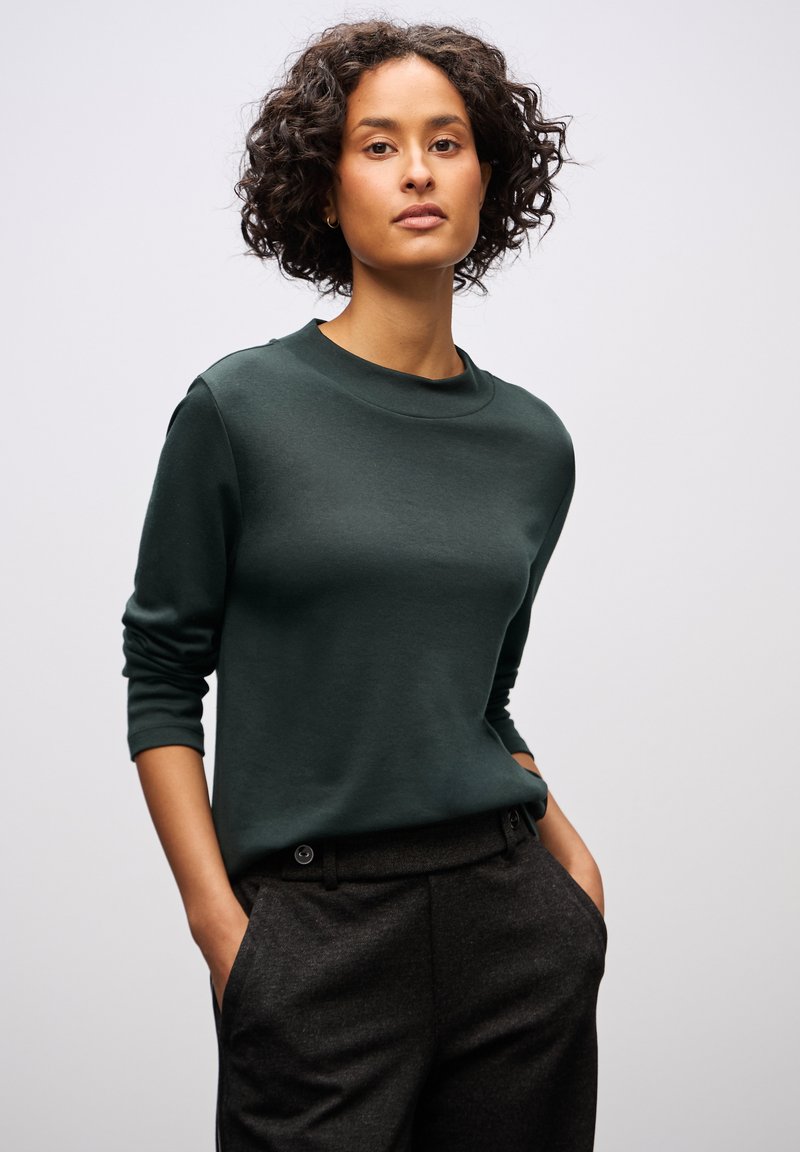 Felpa a girocollo verde scuro con maniche lunghe, texture liscia, silhouette aderente, abbinata a pantaloni grigi scuri con dettagli di bottone.