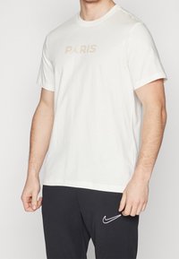 T-shirt en coton blanc avec un graphique beige discret "PARIS" et un logo Jumpman. Manches courtes et encolure ronde.