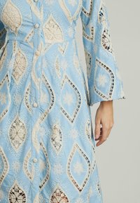 Hellblaues Kleid mit aufwendigen cremefarbenen Spitzenmustern in Diamant- und Blumenmotiven, langen Ärmeln und Knopfdetails.