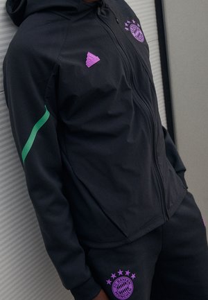Persona vistiendo chándal negro del FC Bayern Múnich con logos morados y una franja verde en la manga, de pie frente a una pared a rayas.