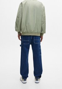 Giubbotto bomber verde con orlo elastico, abbinato a pantaloni cargo in denim scuro larghi con diverse tasche e dettagli di cucitura bianca.