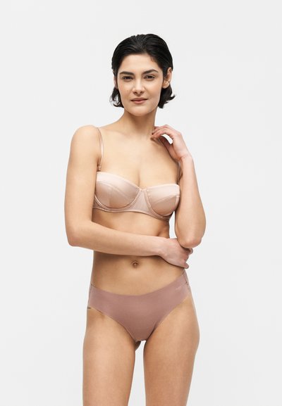 Frau mit kurzen dunklen Haaren, die einen nudefarbenen Balconette-BH und mauve nahtlose hochgeschnittene Unterwäsche trägt, steht mit einer Hand nahe am Gesicht.