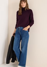 Maglione a collo alto viola con dettagli a coste abbinato a jeans dritti blu chiaro. Una giacca nera è appoggiata su un braccio.