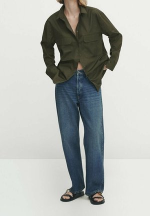 Olive green button-up shirt with two chest pockets, paired with loose blue jeans and black sandals, featuring a relaxed silhouette.
